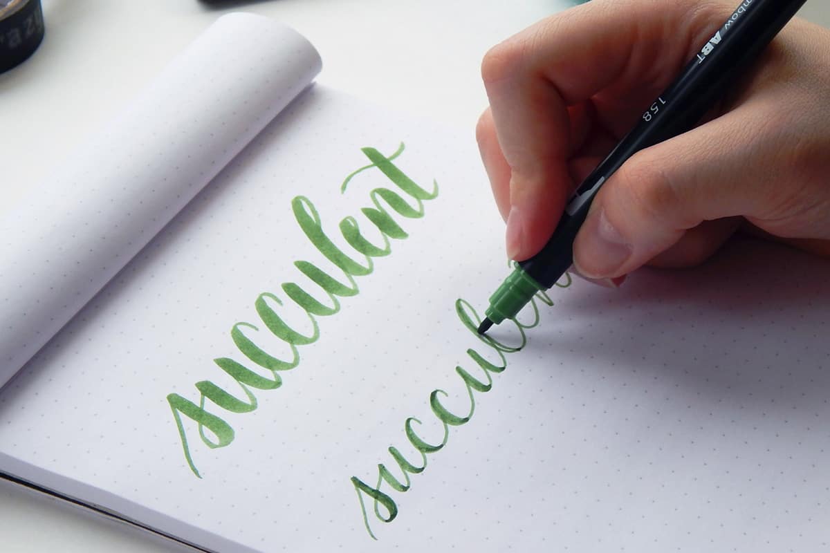 A dot grid notepad is open. A woman uses her right hand and a green Tombow pen to hand letter the word succulent.