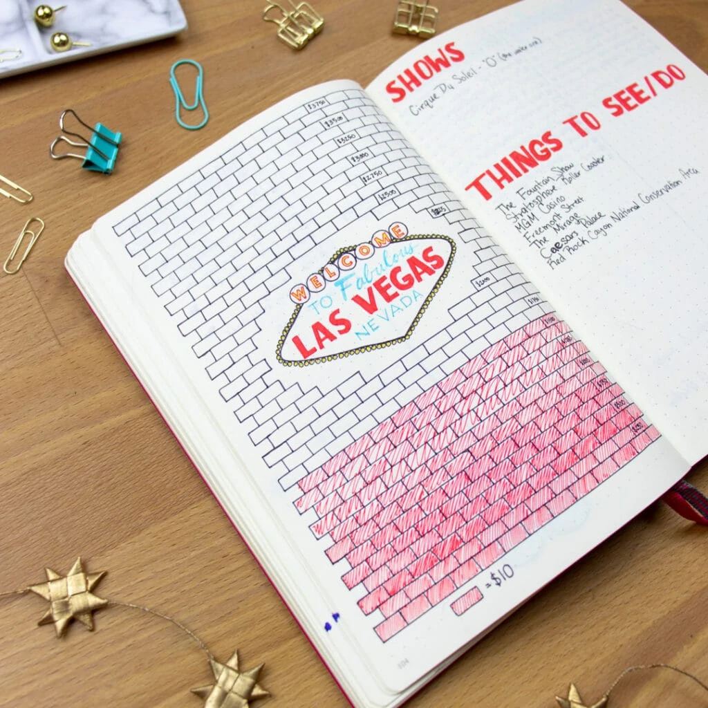 A bullet journal is open to a savings tracker. The tracker is designed like a brick wall, where each brick represents a specific amount of money. The blocks are colored in when money is saved.