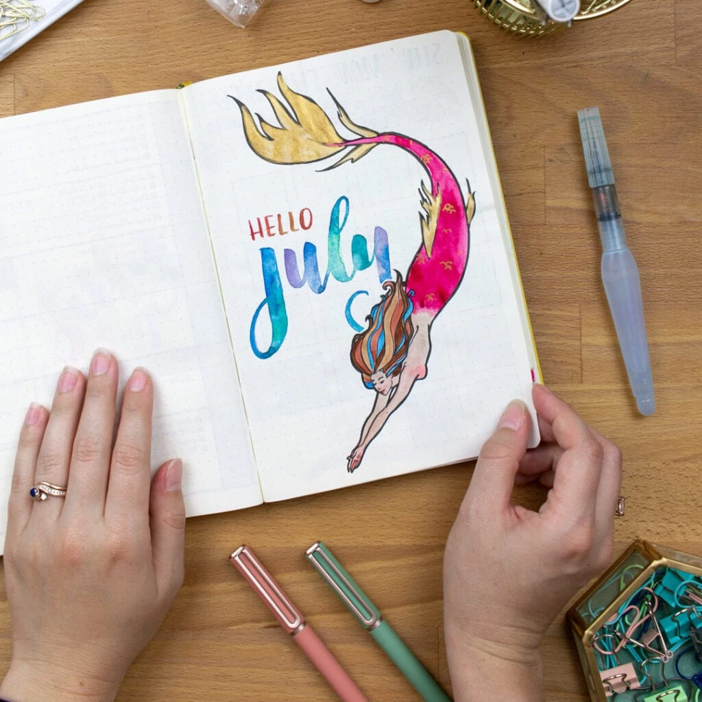 An overhead view of my mermaid bullet journal cover page with a watercolor mermaid and "Hello July" written in watercolor.