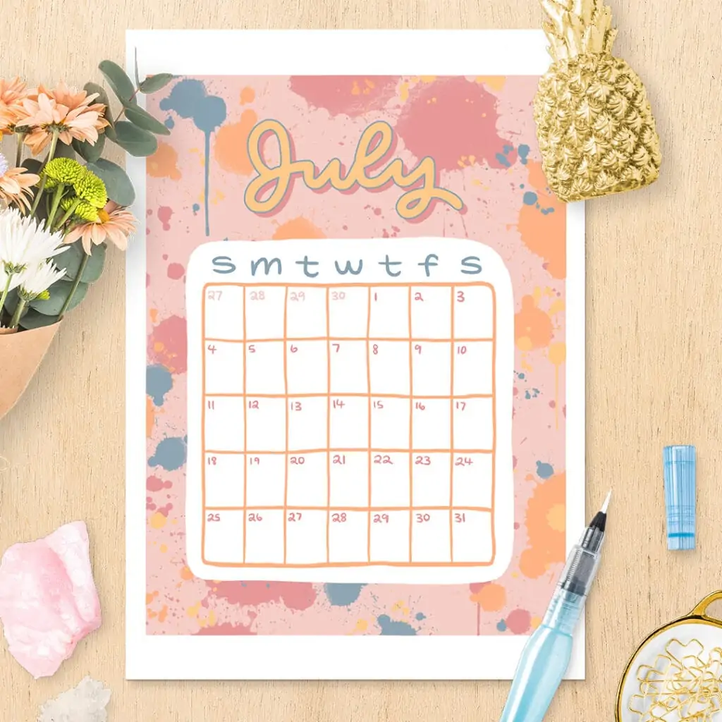 July calendar printable with bright, colorful splash design sitting on wooden desk with flowers, gemstones, and other decor items.