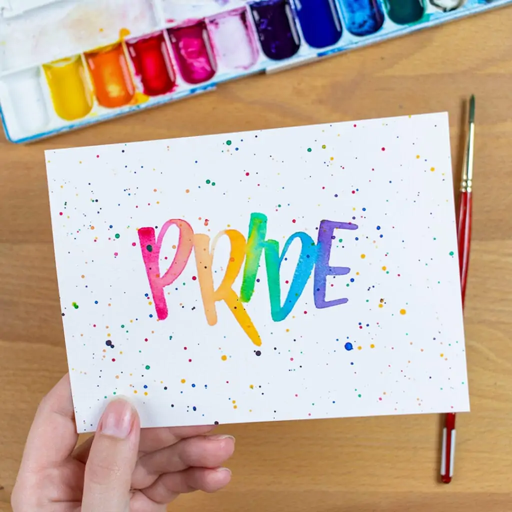 Hand holding small piece of paper that shows "Pride" lettered in rainbow watercolors.
